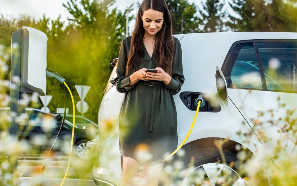 Ragazza Ricarica l Auto Elettrica Vicino ad un Campo di Fiori Ragazza Ricarica l Auto Elettrica Vicino ad un Campo di Fiori